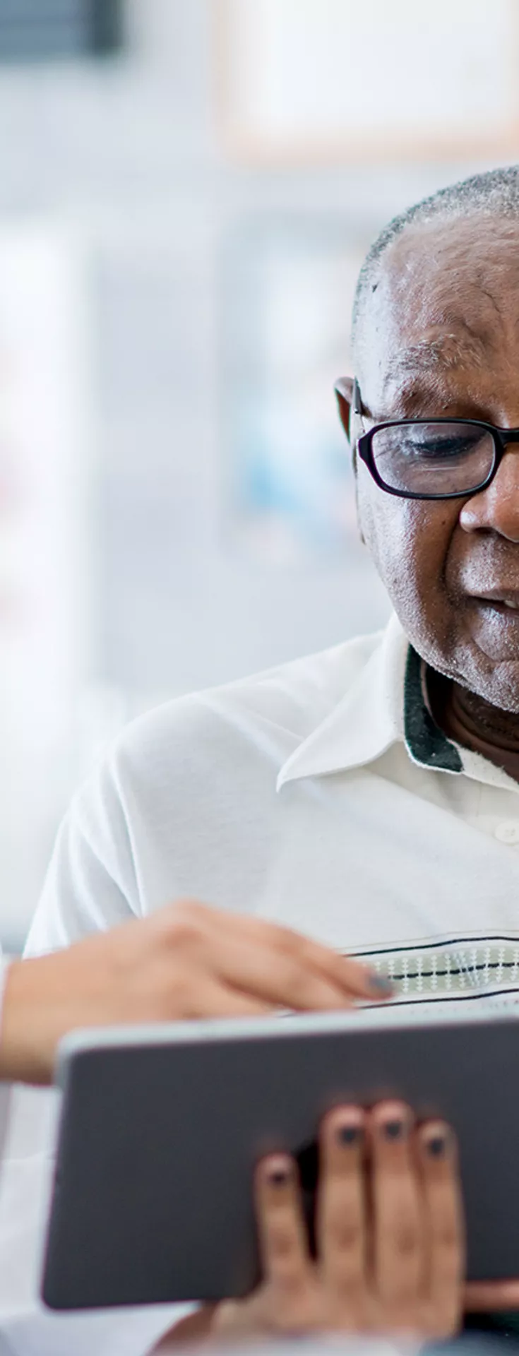 A healthcare professional in a white coat discusses information with an older adult male patient using a digital tablet. The setting appears to be a modern medical office or clinic, with anatomical charts visible in the background. 