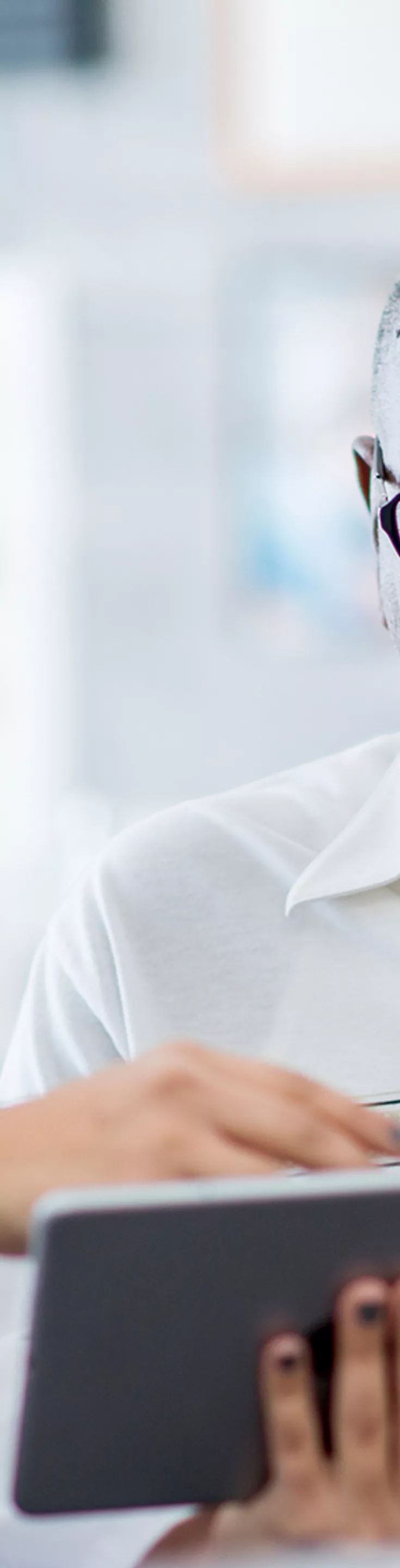 A healthcare professional in a white coat discusses information with an older adult male patient using a digital tablet. The setting appears to be a modern medical office or clinic, with anatomical charts visible in the background. 