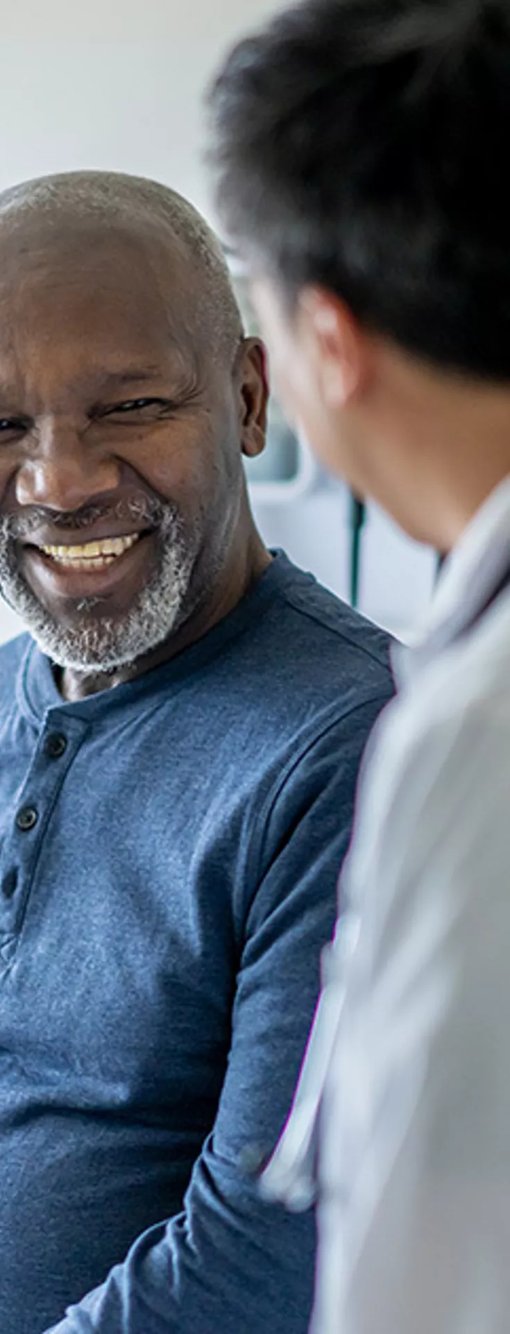 A healthcare professional in a white coat is speaking with a seated adult patient in a modern medical examination room. The setting features bright natural light and clinical decor, emphasizing a professional healthcare environment. The patient is dressed casually in a blue long-sleeve shirt, while the doctor appears attentive and engaged. 