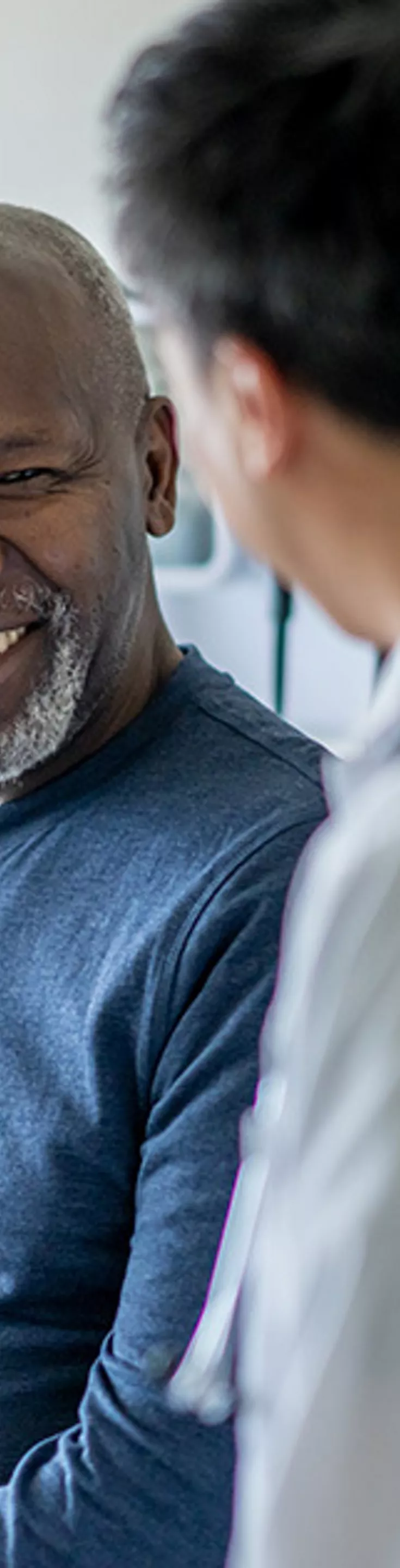 A healthcare professional in a white coat is speaking with a seated adult patient in a modern medical examination room. The setting features bright natural light and clinical decor, emphasizing a professional healthcare environment. The patient is dressed casually in a blue long-sleeve shirt, while the doctor appears attentive and engaged. 
