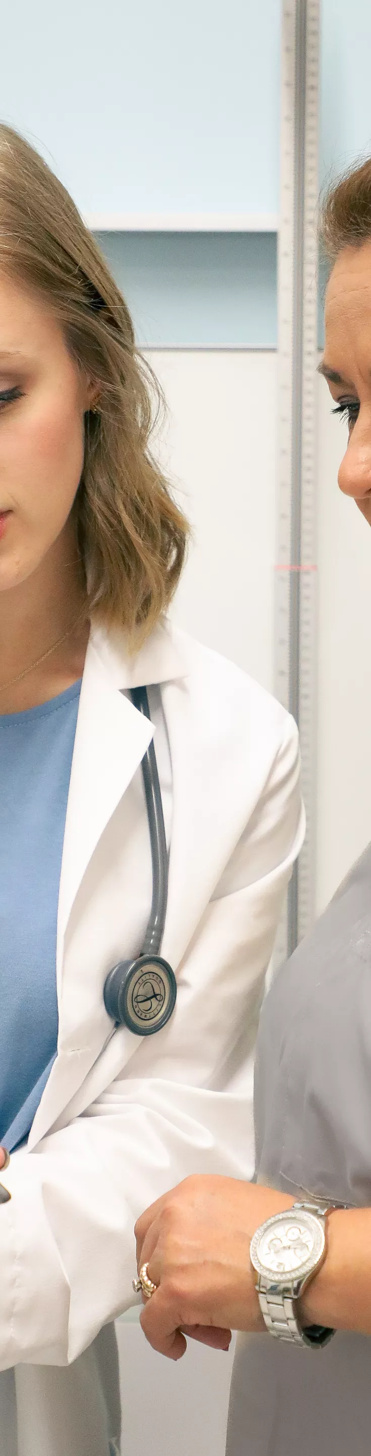 Two female healthcare workers stand together in a clinical setting, one wearing a white coat and the other in gray scrubs. They are focused on a digital tablet, discussing information, with a height measuring device visible in the background. The environment is clean and professional, suggesting a medical or hospital context. 