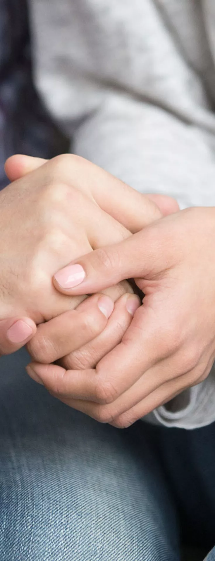 The image shows a close-up of two individuals sitting side by side, holding hands in a gesture of support and reassurance. One person wears a plaid shirt, while the other is dressed in a light gray sweater and blue jeans.
