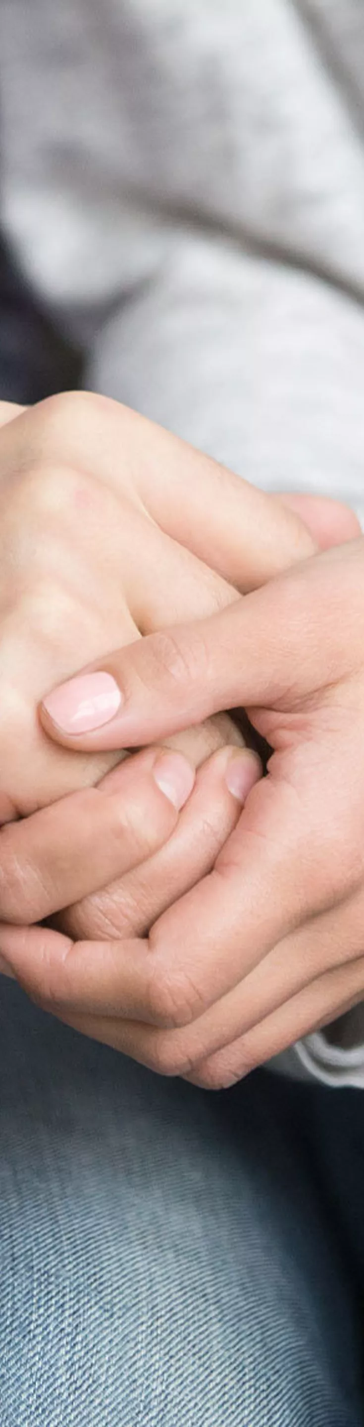 The image shows a close-up of two individuals sitting side by side, holding hands in a gesture of support and reassurance. One person wears a plaid shirt, while the other is dressed in a light gray sweater and blue jeans.