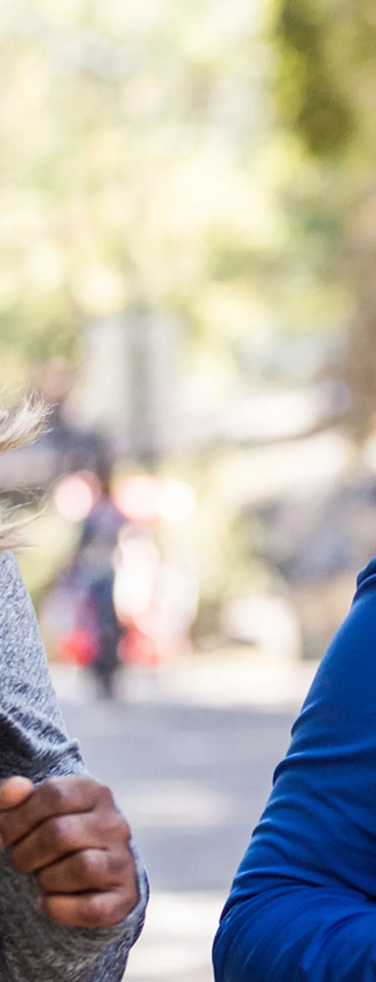 Two women are jogging together along a tree-lined path in a park. They are dressed in athletic wear, one in a gray top and the other in a blue jacket with green accents. The background features blurred trees and other people, suggesting a lively, active environment. 