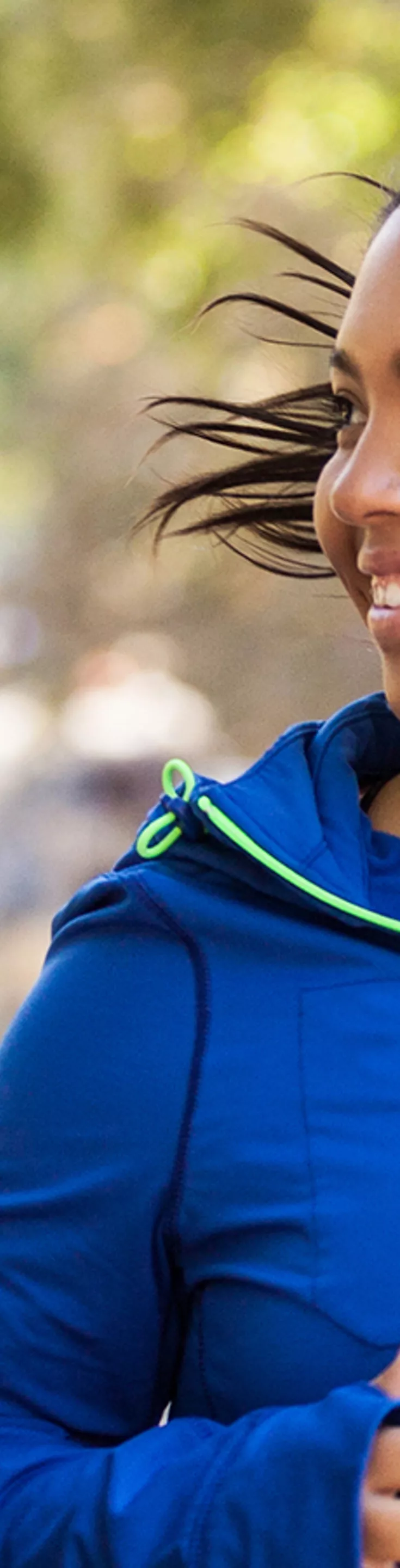 Two women are jogging together along a tree-lined path in a park. They are dressed in athletic wear, one in a gray top and the other in a blue jacket with green accents. The background features blurred trees and other people, suggesting a lively, active environment. 