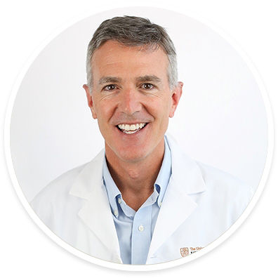 A middle-aged male wearing a white lab coat and light blue collared shirt is pictured against a plain white background. The subject's attire and setting suggest a professional medical or healthcare environment. The lab coat features a small embroidered text on the left side, indicating possible institutional affiliation. The overall image is clean and clinical, emphasizing professionalism.