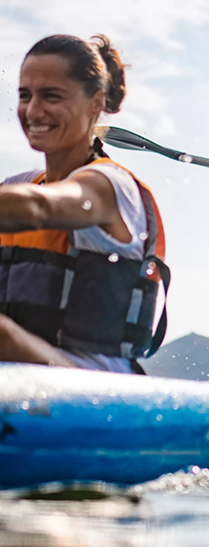 Two individuals paddle a blue kayak together on a sunny day, creating splashes as they move through the water. Both are wearing life vests and casual outdoor clothing, suggesting a recreational activity. The scene captures dynamic movement and teamwork, with a bright and energetic atmosphere. 