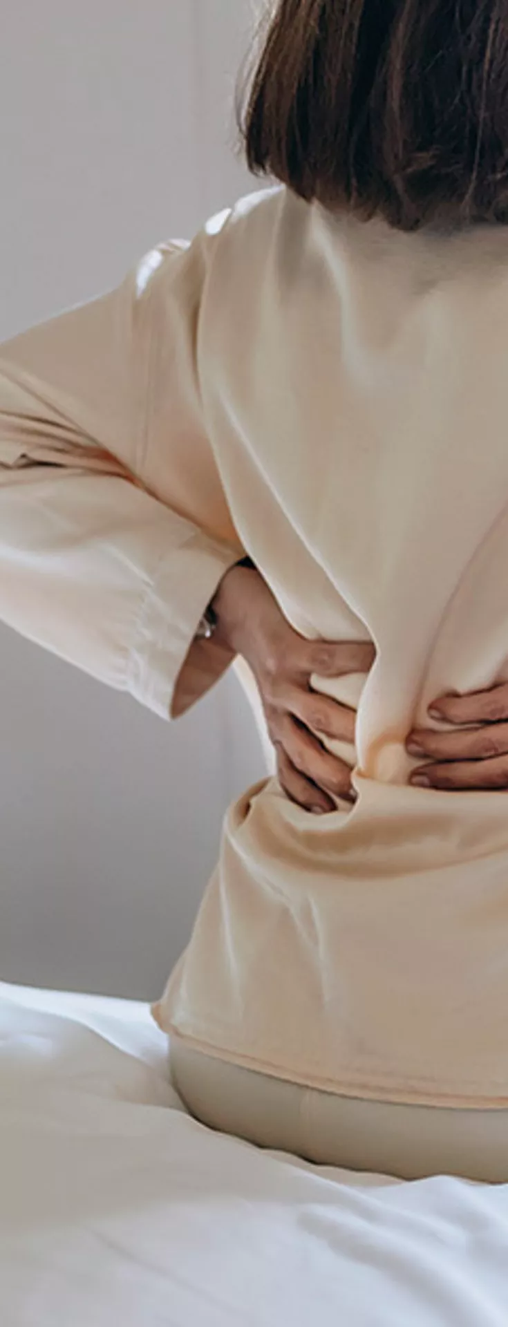 A woman sits on the edge of a bed, facing away, with both hands pressed to her lower back. She appears to be experiencing discomfort or pain, possibly related to her back. The setting is a softly lit bedroom with neutral tones, a window, and a radiator visible. The mood is calm but suggests physical discomfort.