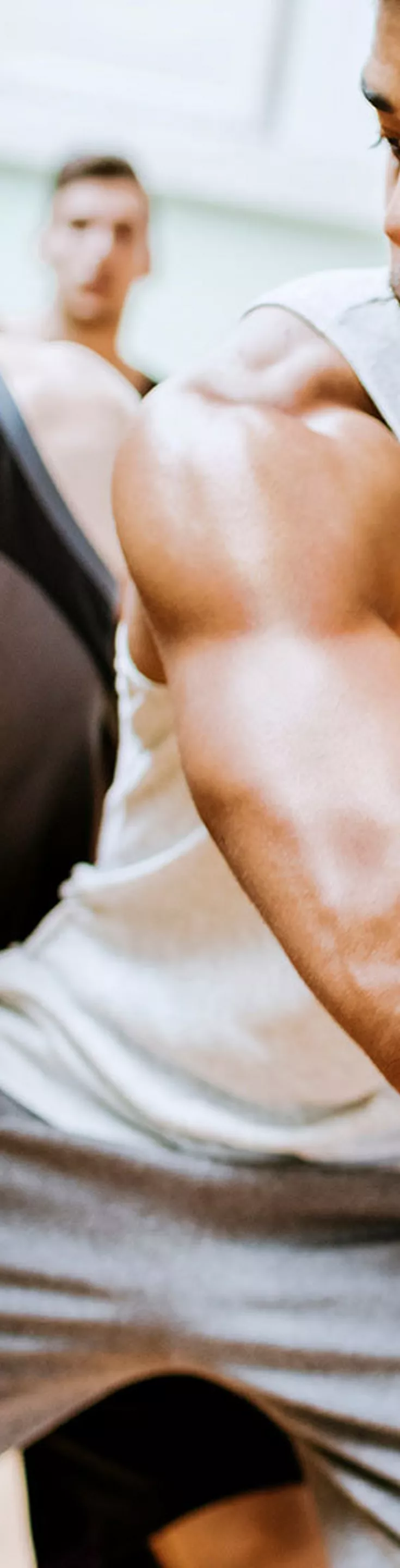 A group of young men are actively playing basketball in an indoor gymnasium. The main subject is holding a basketball, preparing to make a move while being closely guarded by another player. The background shows additional players and a bright, well-lit sports environment. 