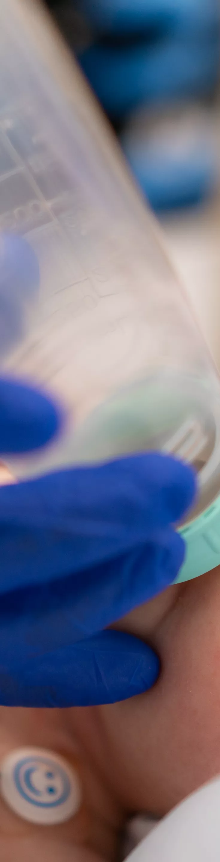 A newborn baby wearing a striped hat and hospital blanket is drinking from a bottle that a gloved medical professional holds over the infant's face. The setting appears to be a hospital or medical facility, with the baby lying on a bed. 