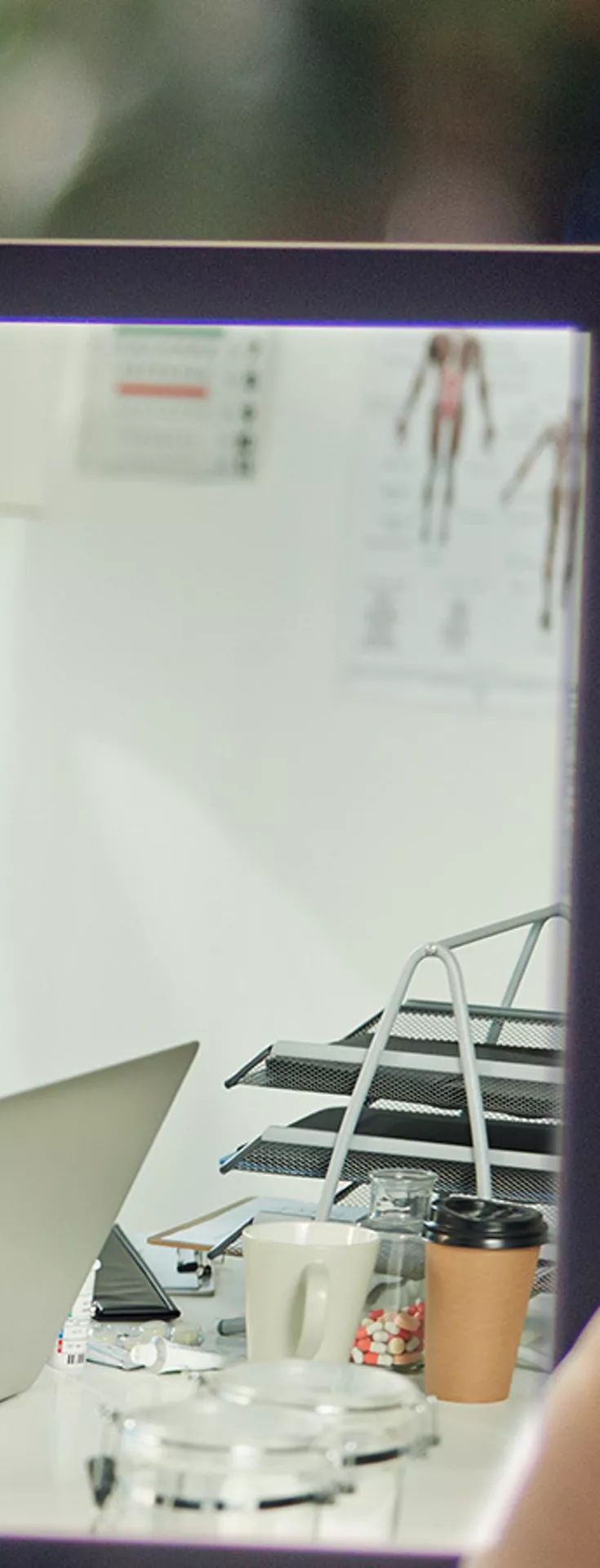 A person is holding a tablet displaying a video call with a healthcare professional. The medical expert is wearing blue scrubs and appears to be explaining something, possibly related to health or treatment. The setting suggests a home environment, with the tablet screen showing a desk, laptop, and medical equipment. 