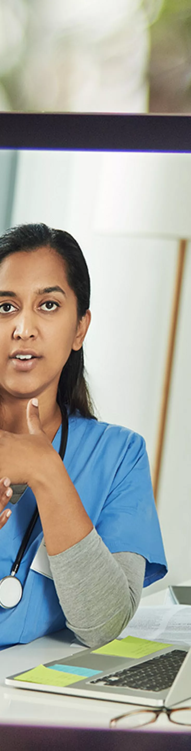 A person is holding a tablet displaying a video call with a healthcare professional. The medical expert is wearing blue scrubs and appears to be explaining something, possibly related to health or treatment. The setting suggests a home environment, with the tablet screen showing a desk, laptop, and medical equipment. 