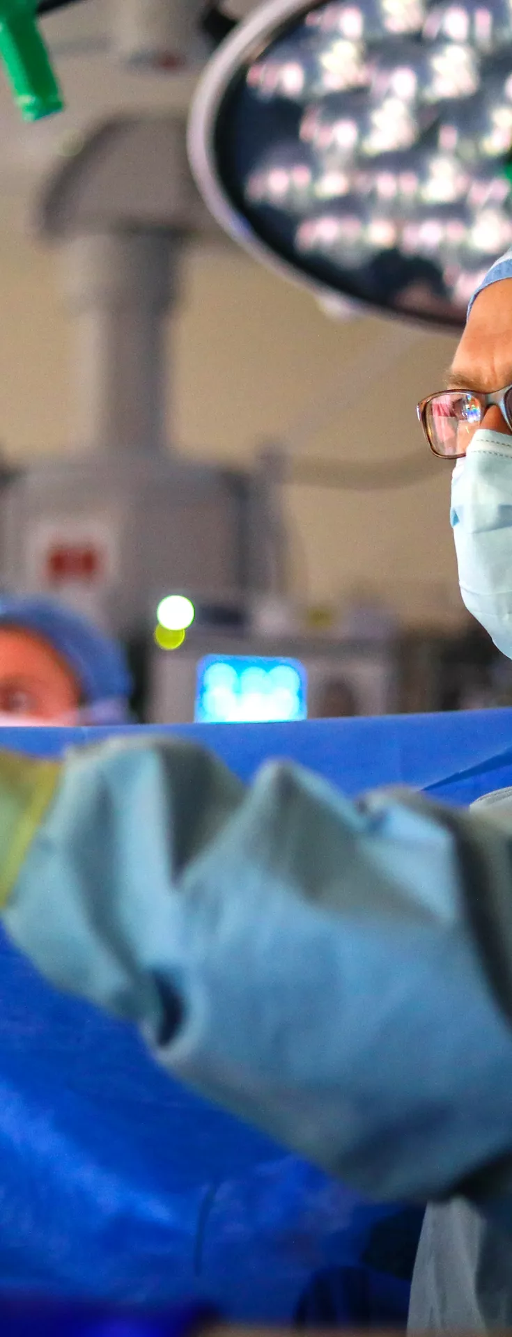 A medical professional in surgical attire is seen working in a brightly lit operating room. The individual is wearing a blue gown, gloves, and a surgical cap, surrounded by advanced medical equipment and monitors. The environment is sterile and clinical, with overhead surgical lights illuminating the workspace. 