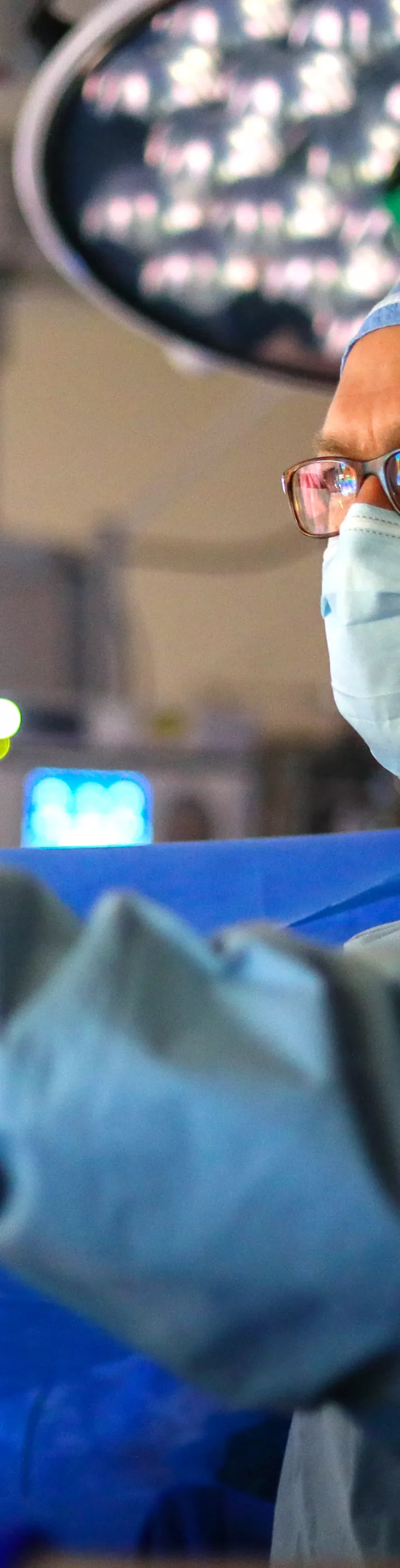 A medical professional in surgical attire is seen working in a brightly lit operating room. The individual is wearing a blue gown, gloves, and a surgical cap, surrounded by advanced medical equipment and monitors. The environment is sterile and clinical, with overhead surgical lights illuminating the workspace. 