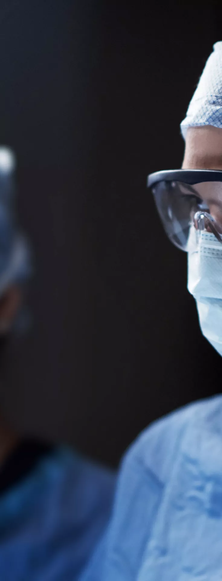 Two medical professionals are shown in an operating room environment, dressed in blue surgical gowns, caps, and masks. The background is dimly lit, emphasizing the sterile and focused atmosphere typical of surgical procedures. 