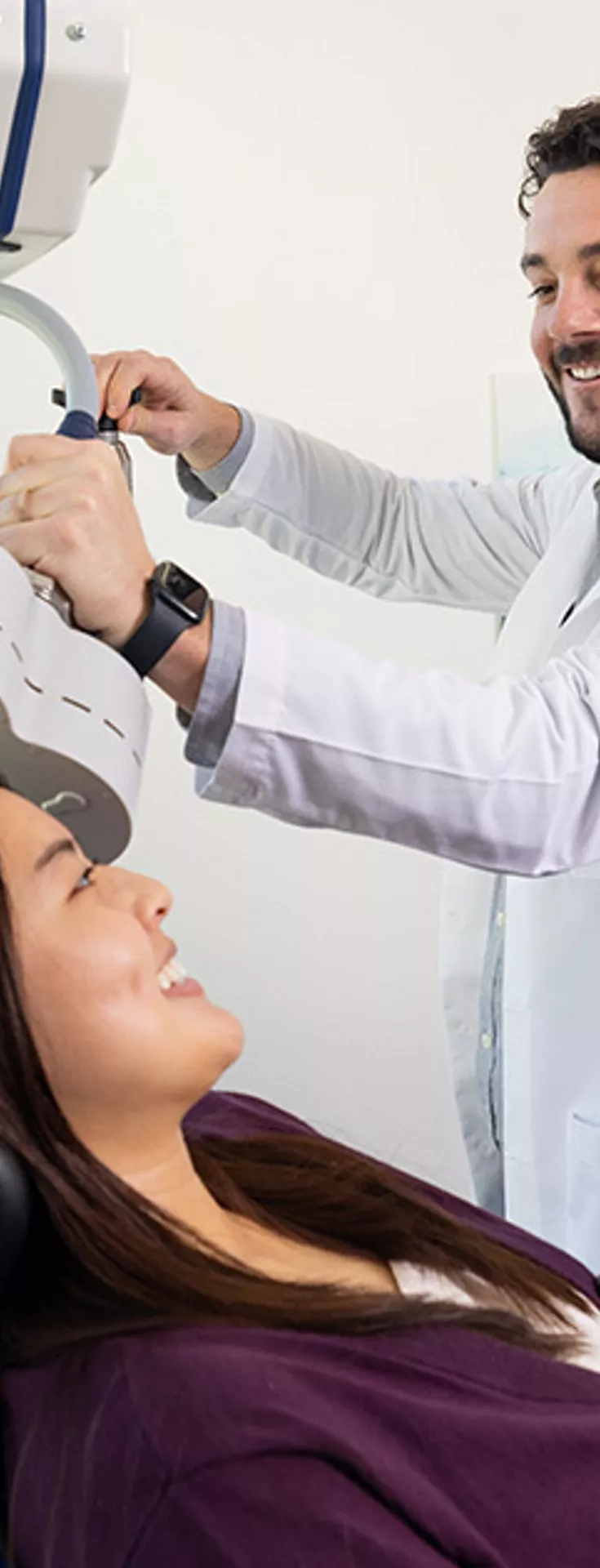 A healthcare professional operates a specialized medical device on a seated patient in a modern clinical setting. The room features large windows with an urban view and contemporary decor. The doctor is wearing a white lab coat, and the patient is positioned comfortably for the procedure. 