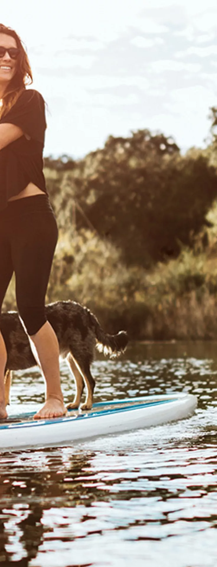 A woman stands on a paddleboard, paddling across a tranquil lake with her dog. The scene is set outdoors in nature, surrounded by lush trees and bathed in warm, golden sunlight. Both the woman and the dog appear relaxed, enjoying the peaceful water and scenic environment.