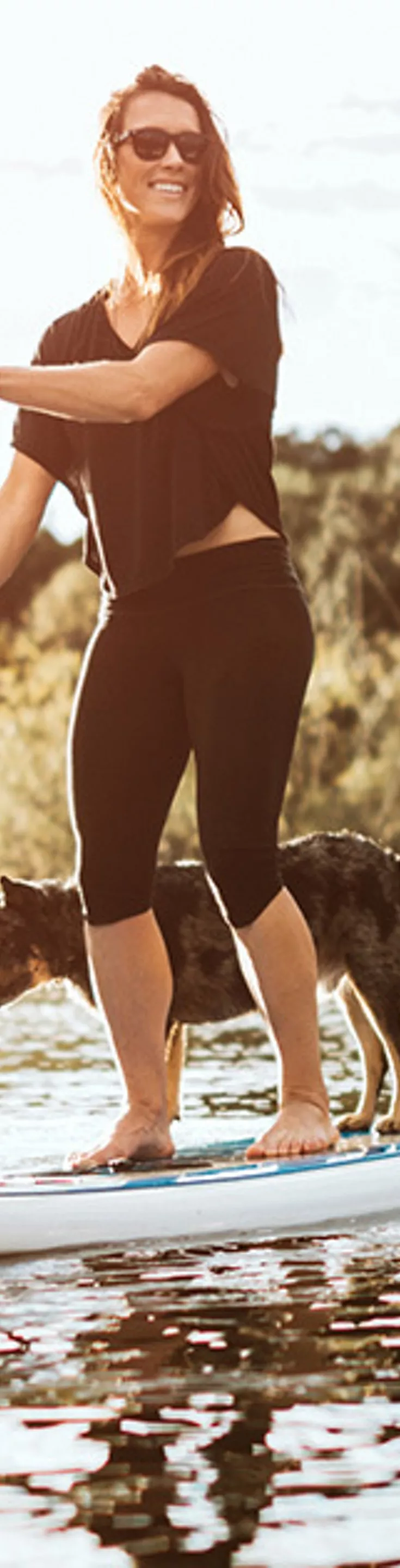 A woman stands on a paddleboard, paddling across a tranquil lake with her dog. The scene is set outdoors in nature, surrounded by lush trees and bathed in warm, golden sunlight. Both the woman and the dog appear relaxed, enjoying the peaceful water and scenic environment.