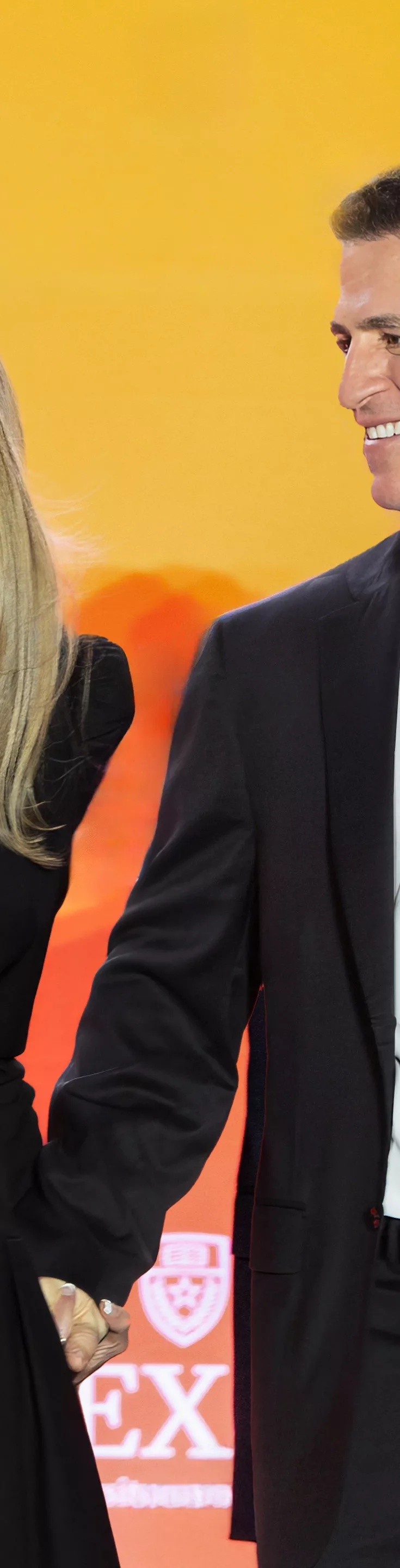 Michael and Susan Dell at the What Starts Here Event on April 21st, 2026. They are dressed in formal black attire, holding hands on stage, and are looking off to their right. The background is a bright yellow and orange digital display with partial text visible - "What starts here" and "Texas" are implied.