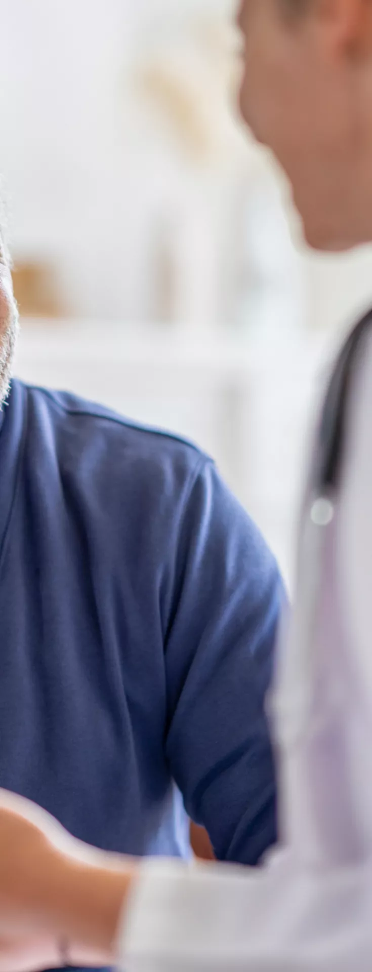 An older man sits across from a healthcare worker in a bright, indoor setting. The man is wearing a blue cardigan and shirt, while the professional is dressed in white attire, possibly a nurse or doctor. They appear to be engaged in a discussion, with the man holding a tablet or clipboard. 