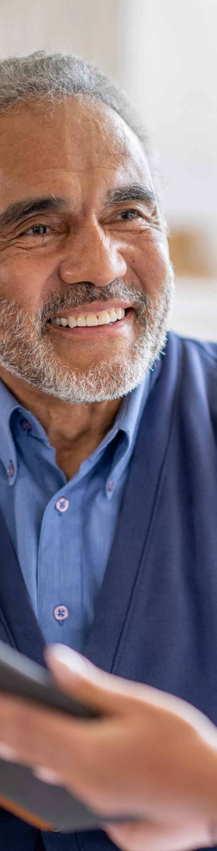 An older man sits across from a healthcare worker in a bright, indoor setting. The man is wearing a blue cardigan and shirt, while the professional is dressed in white attire, possibly a nurse or doctor. They appear to be engaged in a discussion, with the man holding a tablet or clipboard. 