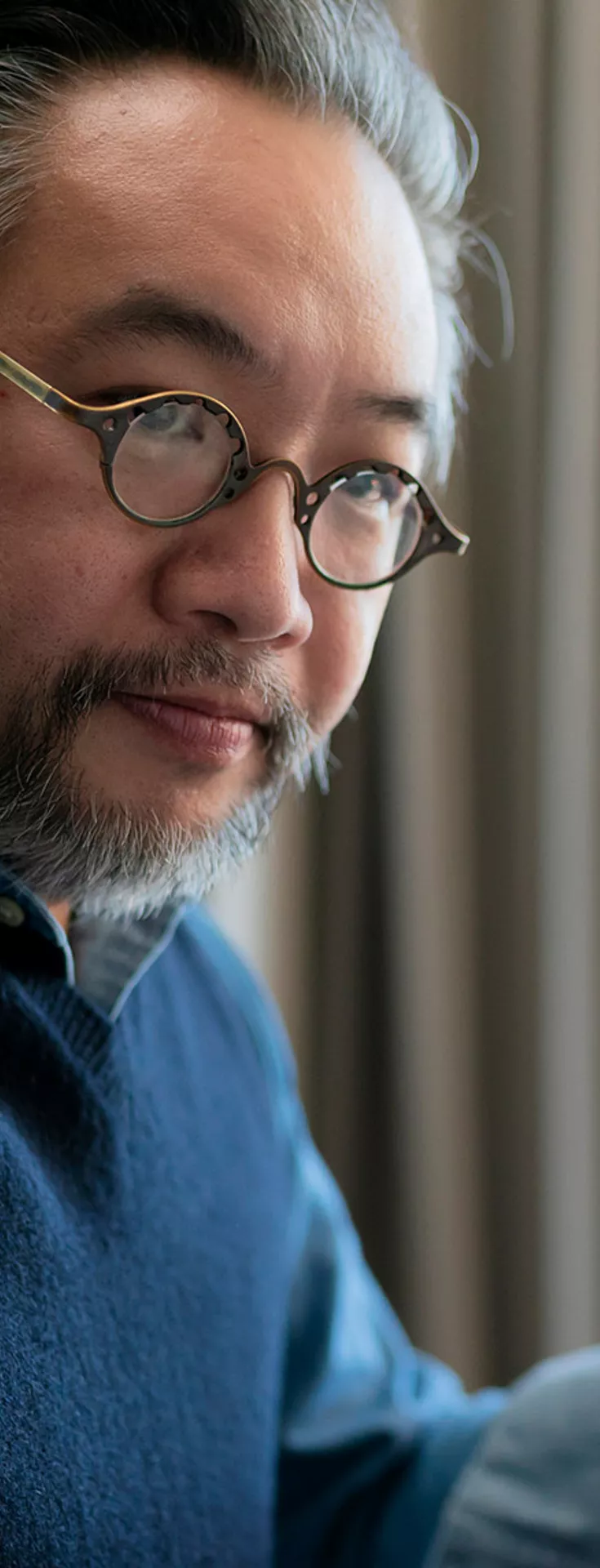 A person is seated at a desk, holding an orange mug in one hand. The background features beige curtains and a modern desk lamp, suggesting a cozy home office or study environment. The individual is wearing a blue sweater vest over a light shirt, and the scene is softly lit with natural daylight. 