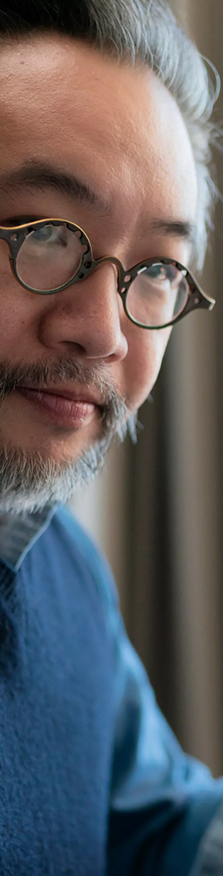 A person is seated at a desk, holding an orange mug in one hand. The background features beige curtains and a modern desk lamp, suggesting a cozy home office or study environment. The individual is wearing a blue sweater vest over a light shirt, and the scene is softly lit with natural daylight. 