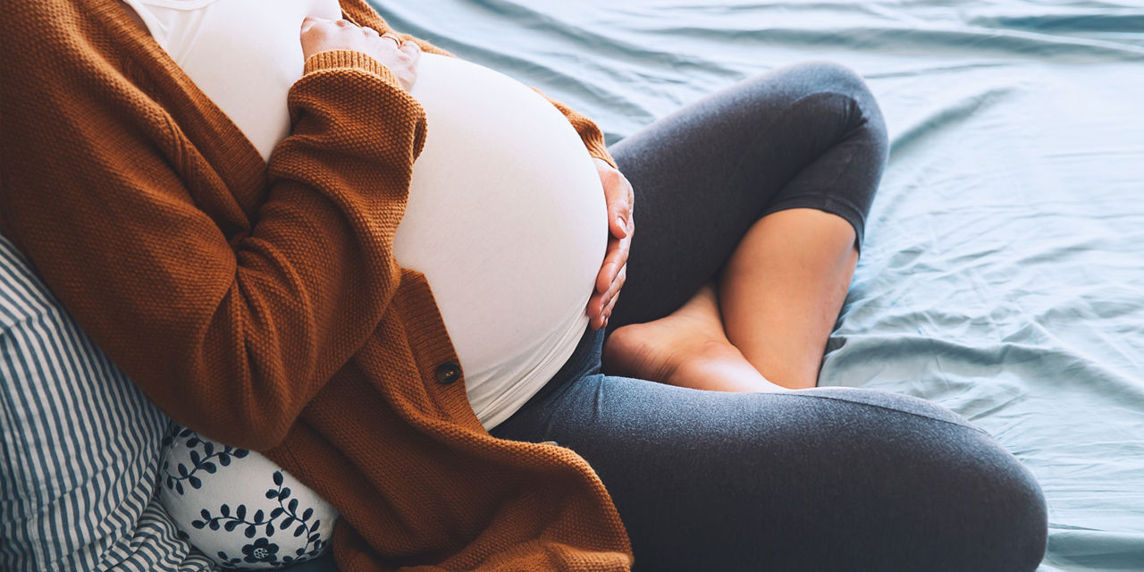 A pregnant woman sits cross-legged on a bed, gently holding her belly. She is dressed in a white top, brown cardigan, and dark leggings, with a patterned pillow supporting her back. The scene conveys comfort and calmness, with soft natural lighting and a relaxed atmosphere.