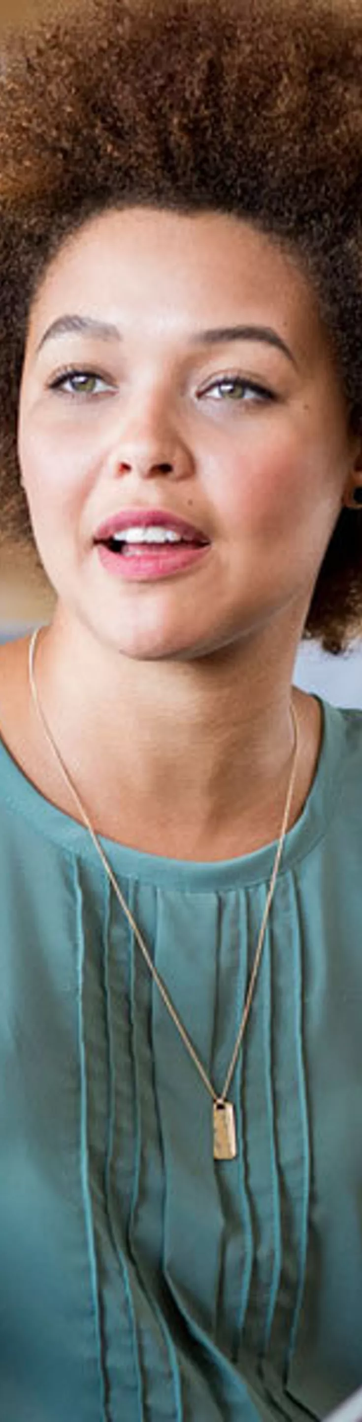 Two women are engaged in conversation while seated on a comfortable gray sofa in a contemporary lounge or office space. One woman gestures expressively with her hands, while the other holds a tablet device. The background features framed pictures on a wooden wall, contributing to a casual and collaborative atmosphere.