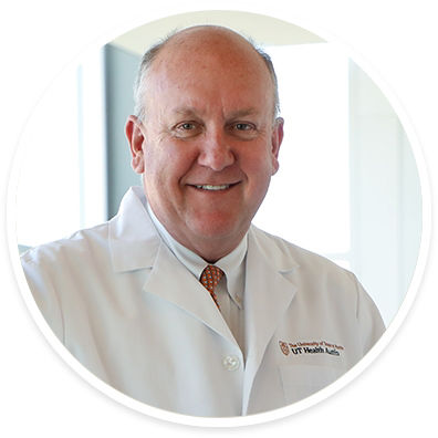 An older male doctor is pictured wearing a white lab coat with a visible UT Health Austin insignia. He stands indoors in a well-lit, professional medical environment. The doctor's attire includes a white shirt and patterned tie, suggesting a formal clinical setting. The image conveys professionalism and expertise in healthcare.