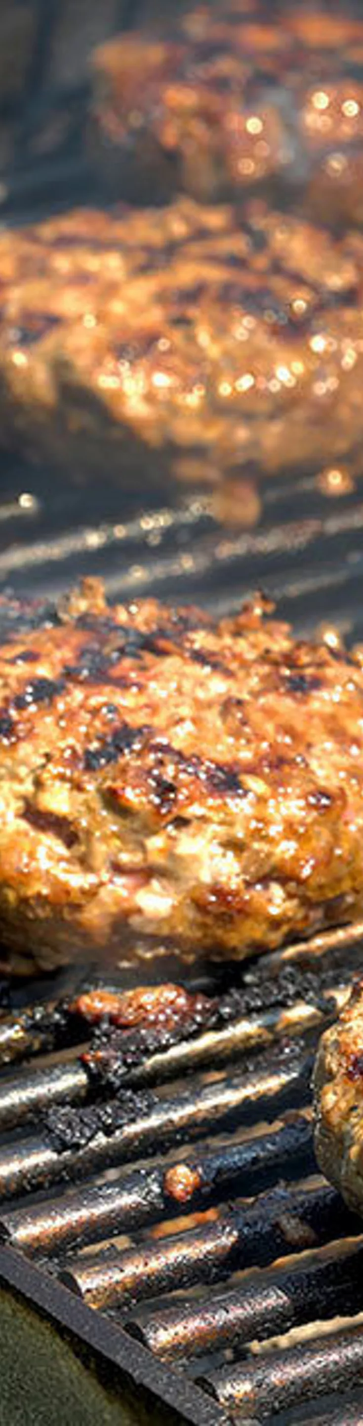 Several hamburger patties and hot dogs are cooking on a metal grill, with visible grill marks and smoke rising. The scene suggests an outdoor barbecue, likely during a casual gathering or picnic. The food is arranged closely together, highlighting the classic summer cookout atmosphere. No visible text, numbers, or brand markings are present in the image.