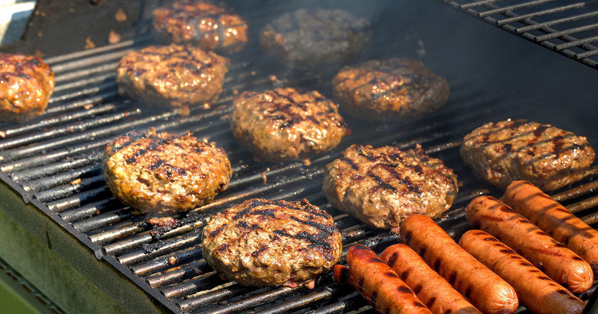 Several hamburger patties and hot dogs are cooking on a metal grill, with visible grill marks and smoke rising. The scene suggests an outdoor barbecue, likely during a casual gathering or picnic. The food is arranged closely together, highlighting the classic summer cookout atmosphere. No visible text, numbers, or brand markings are present in the image.