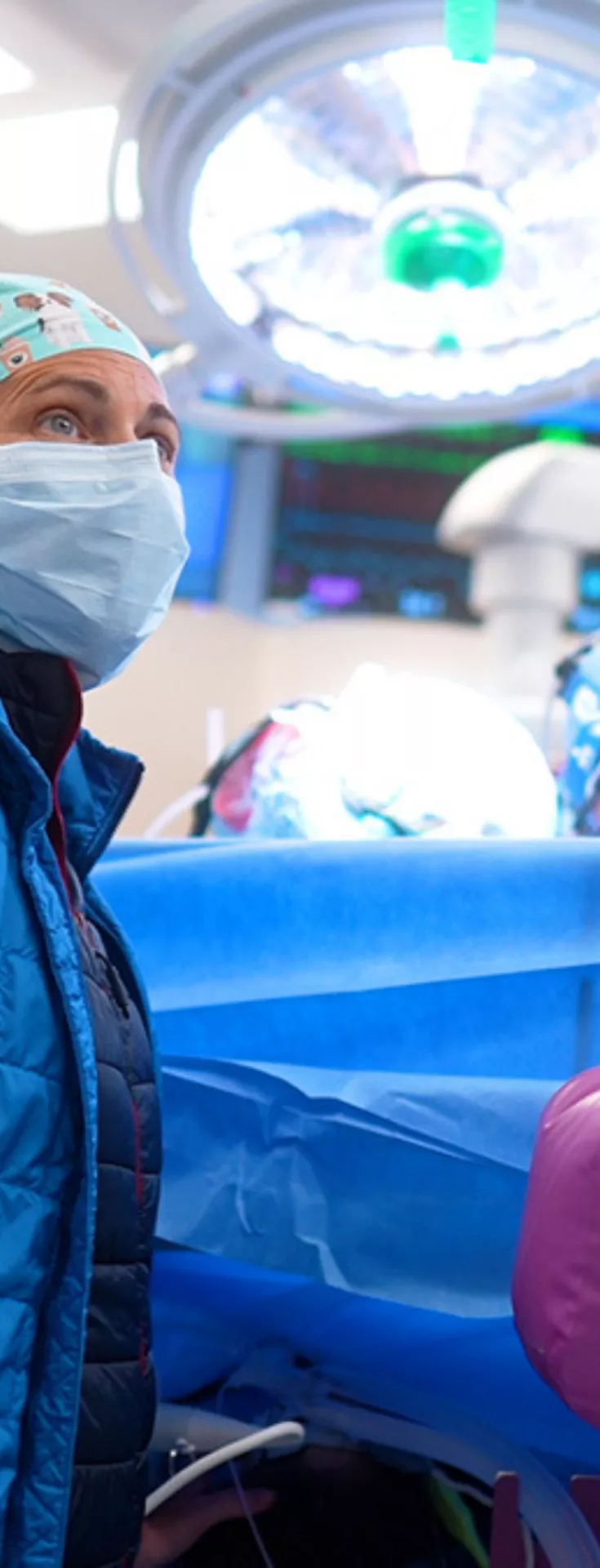 A female surgical nurse, masked with a blue coat monitors a patient, while a surgeon is working on the other side of the draped patient.