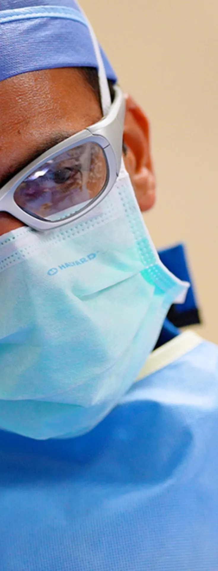 A male surgeon, masked with protective eyewear, tilts his head and looks at a monitor off screen.