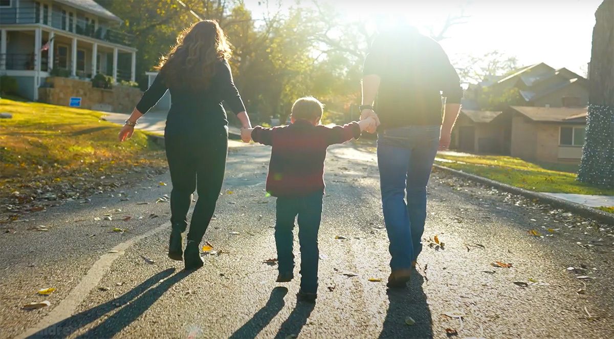 A child walks between two adults, holding their hands, as they stroll down a sunlit residential street. The scene is backlit with warm sunlight, casting long shadows on the pavement. The setting appears to be a quiet suburban neighborhood with houses and trees visible. The mood is joyful and relaxed, emphasizing togetherness and family bonding. No visible text or numbers are present in the image.