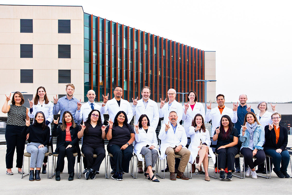 A large group of adults, including men and women, pose together in front of a modern building. Several individuals are wearing white lab coats, suggesting a medical or scientific team, while others are dressed in business casual attire. The group is seated and standing, smiling and making hand gestures, with a contemporary glass and concrete building in the background. The setting appears to be outdoors, possibly on a rooftop or terrace. The overall mood is professional and friendly.