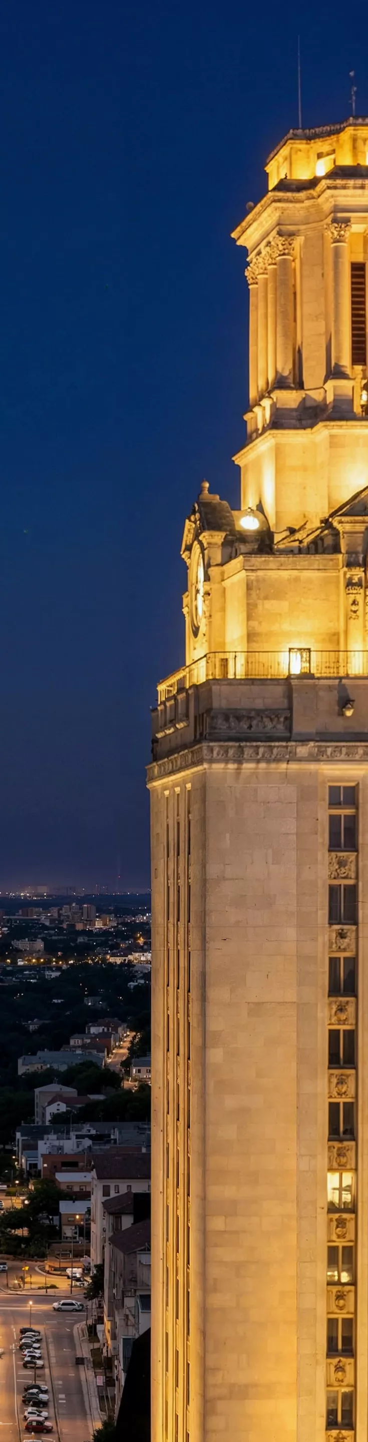 The UT Austin tower stands brightly lit against a deep blue evening sky, overlooking a sprawling urban landscape with a bright tower in the background. 