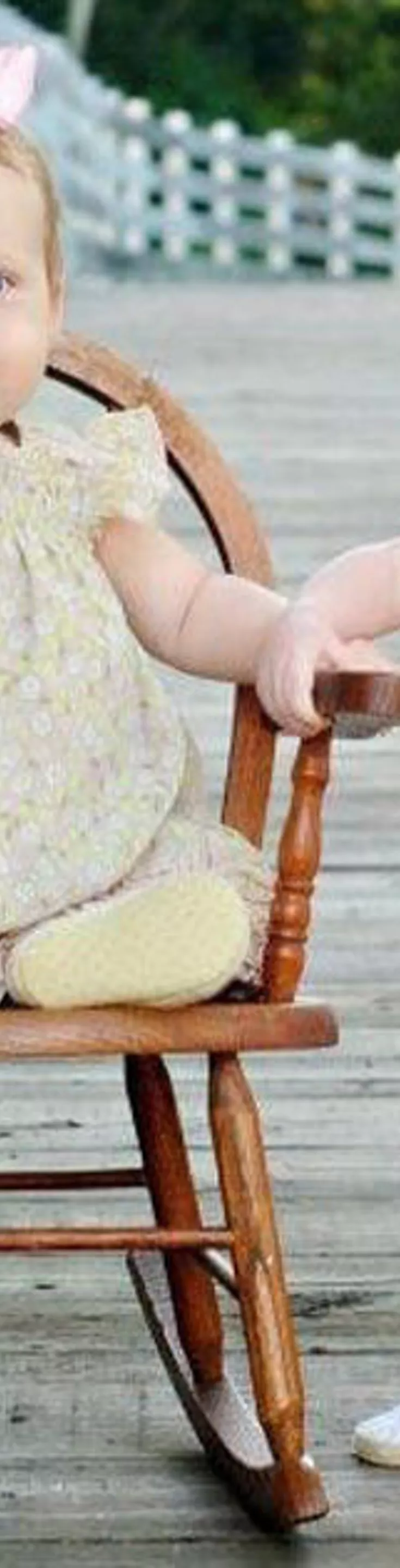 Two small children are featured on a wooden bridge, one sitting in a rocking chair and the other standing beside her. Both girls are wearing pastel floral dresses with matching bows in their hair. The setting is outdoors, with a rustic wooden railing and greenery in the background. The overall mood is playful and serene, with soft, natural lighting.