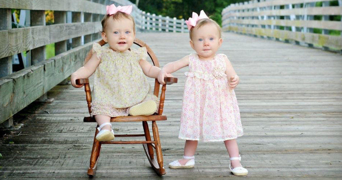 Two small children are featured on a wooden bridge, one sitting in a rocking chair and the other standing beside her. Both girls are wearing pastel floral dresses with matching bows in their hair. The setting is outdoors, with a rustic wooden railing and greenery in the background. The overall mood is playful and serene, with soft, natural lighting.
