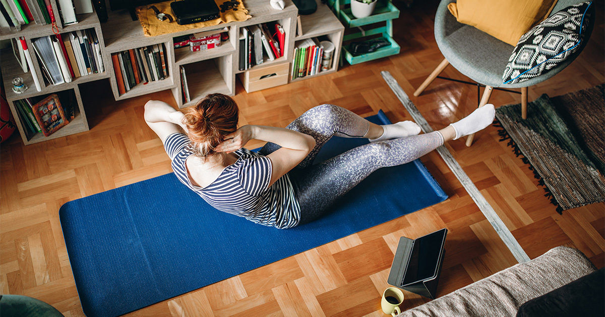 A young woman performs a core workout on a blue yoga mat in a well-lit living room. She is wearing striped activewear and is surrounded by bookshelves, a tablet, and home decor. The setting is casual and inviting, with wooden flooring and a patterned chair visible. The atmosphere suggests a comfortable home workout routine.