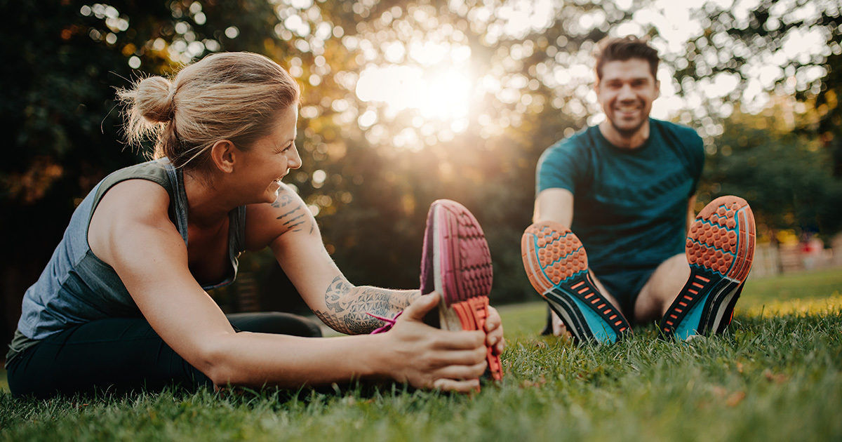 Two adults are stretching on a grassy field during daylight, with the sun shining through the trees in the background. Both individuals are wearing athletic clothing and brightly colored running shoes. The setting suggests a park or outdoor recreational area, and the mood is energetic and healthy. The image captures a moment of physical activity and wellness.