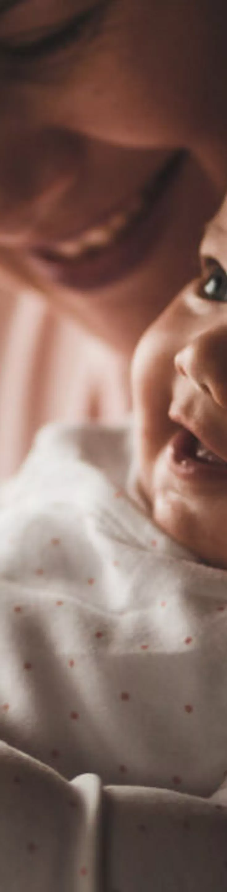 An adult gently cradles a baby, focusing on the baby's feet and hands. The setting appears to be indoors with warm, soft lighting, creating a calm and intimate atmosphere. The baby is dressed in a light-colored, long-sleeve onesie. The image conveys tenderness and care, with a neutral color palette.