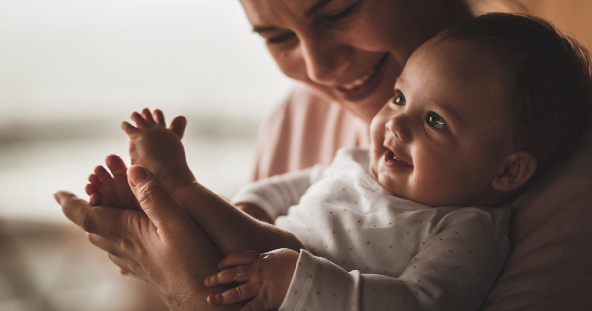 An adult gently cradles a baby, focusing on the baby's feet and hands. The setting appears to be indoors with warm, soft lighting, creating a calm and intimate atmosphere. The baby is dressed in a light-colored, long-sleeve onesie. The image conveys tenderness and care, with a neutral color palette.
