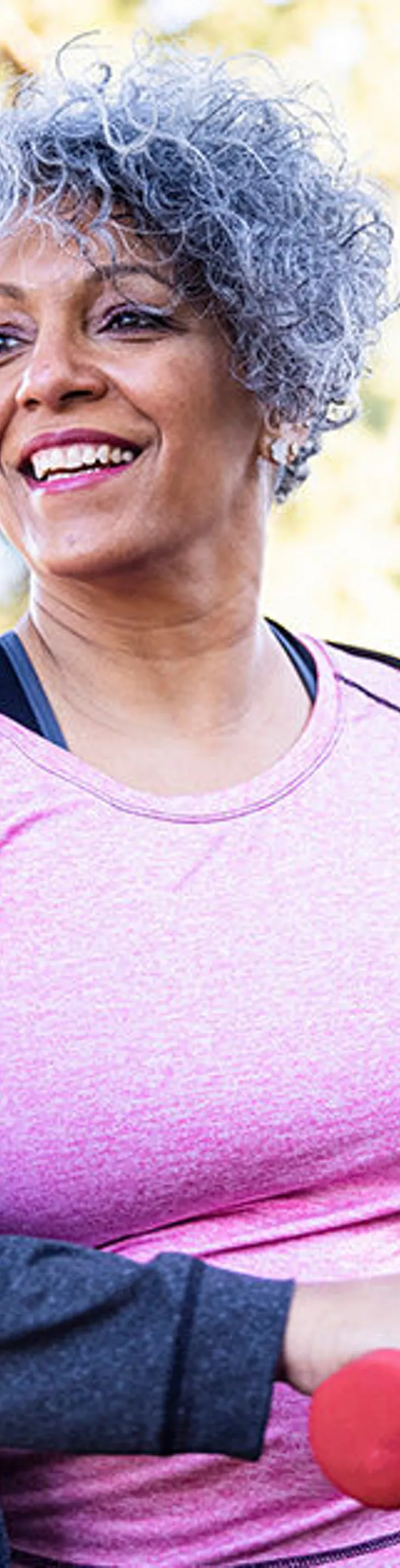 An older woman with short curly gray hair is seen exercising in a park setting, holding small red dumbbells. She wears a pink athletic top and a dark jacket, suggesting a casual fitness routine. The background features trees and sunlight, creating a bright and energetic atmosphere. The image conveys health, vitality, and an active lifestyle.