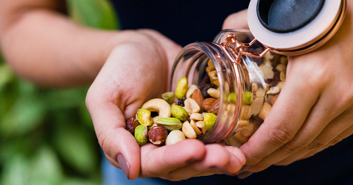 A close-up view shows a person pouring a variety of mixed nuts from a glass jar into their hand. The scene is set outdoors with a blurred green background, suggesting a natural or garden environment. The image highlights the textures and colors of the nuts, including cashews, pistachios, and hazelnuts. The hands appear to belong to an adult, and the overall mood is casual and healthy.