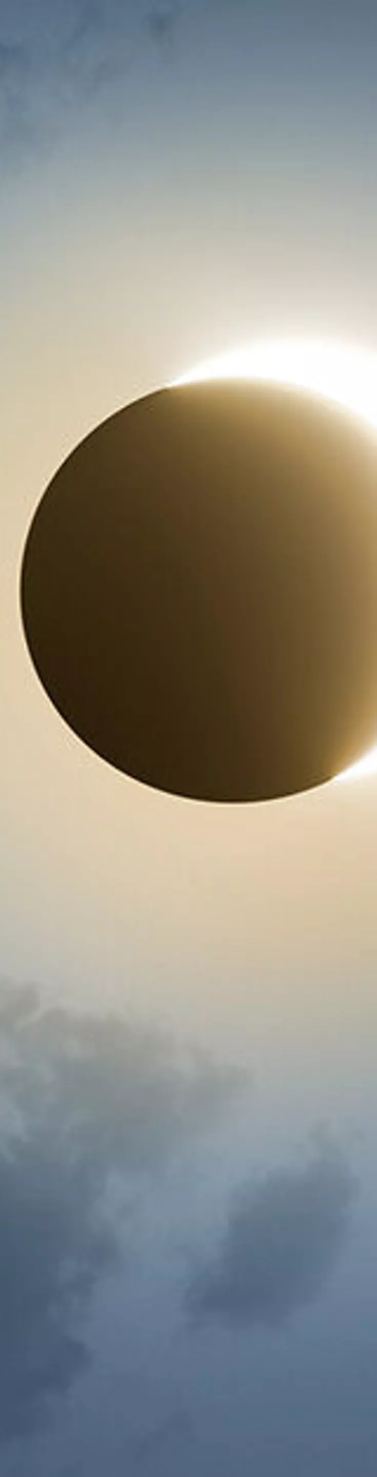 A striking solar eclipse is captured as the moon passes directly in front of the sun, creating a glowing corona effect. Wispy clouds partially obscure the sky, adding depth and atmosphere to the celestial event. The scene is bathed in a cool, ethereal light, emphasizing the contrast between the darkened moon and the bright solar halo. No visible text or numbers are present in the image.