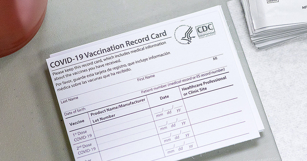 A stack of COVID-19 Vaccination Record Cards is shown on a light-colored table. The card displays fields for vaccine type, product name, manufacturer, lot number, and healthcare professional or clinic site. The CDC logo is clearly visible in the upper right corner. The card includes sections for first and second doses, with date fields formatted as mm/dd/yy. The setting appears clinical and organized, with a neutral color palette.
