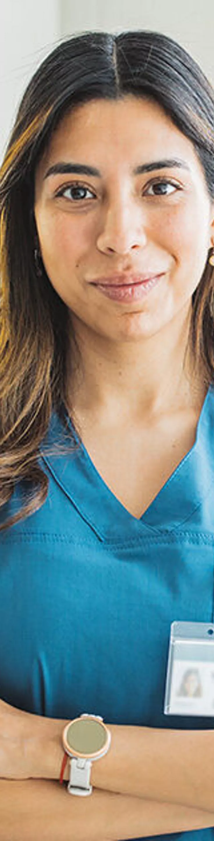 A healthcare worker is shown standing with arms crossed, wearing blue medical scrubs and an identification badge. The setting appears to be a bright, modern medical facility with large windows and a clean, organized environment. The individual is accessorized with a smartwatch and a simple bracelet. The overall mood is professional and calm, with a neutral color palette dominated by blue and white tones.