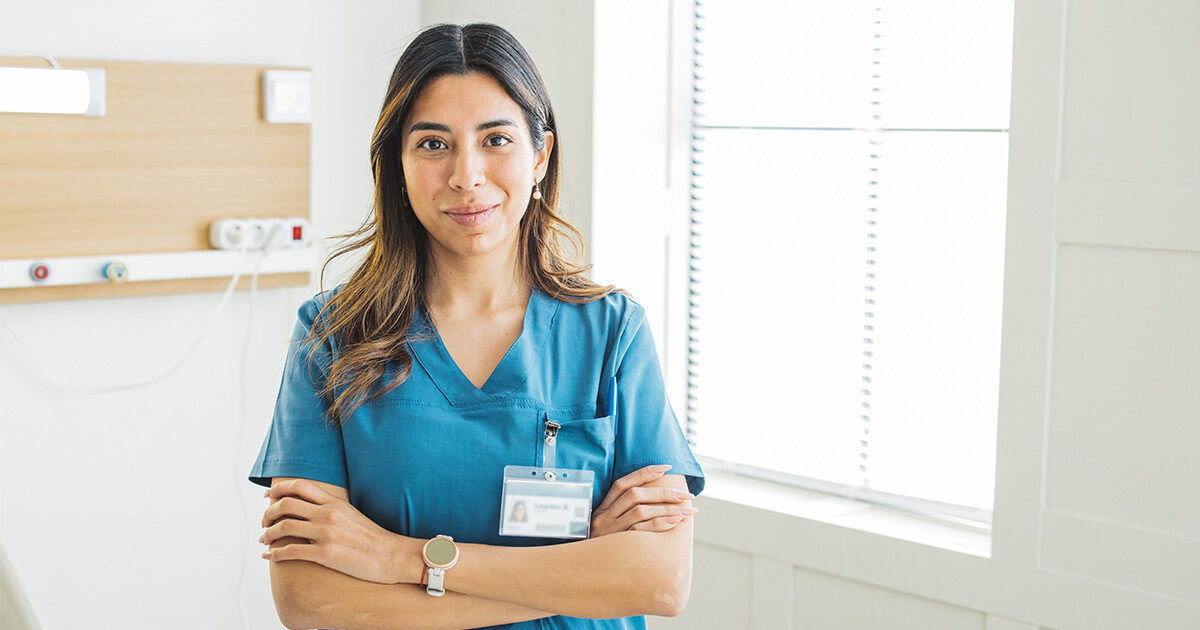 A healthcare worker is shown standing with arms crossed, wearing blue medical scrubs and an identification badge. The setting appears to be a bright, modern medical facility with large windows and a clean, organized environment. The individual is accessorized with a smartwatch and a simple bracelet. The overall mood is professional and calm, with a neutral color palette dominated by blue and white tones.
