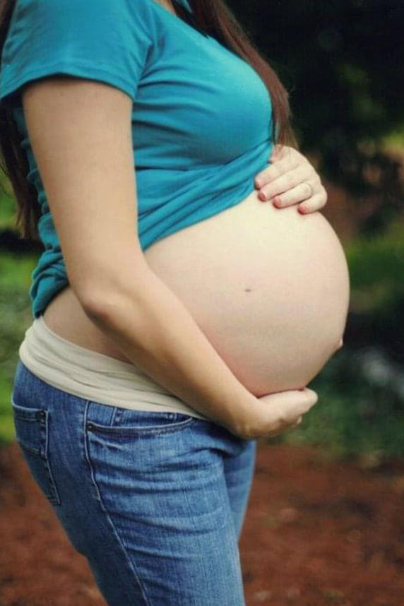 A pregnant woman is shown gently holding her bare belly with both hands. She is wearing a teal shirt and blue jeans, standing in an outdoor setting with greenery and earthy tones in the background. The image conveys a calm, nurturing mood and focuses on the anticipation of motherhood.